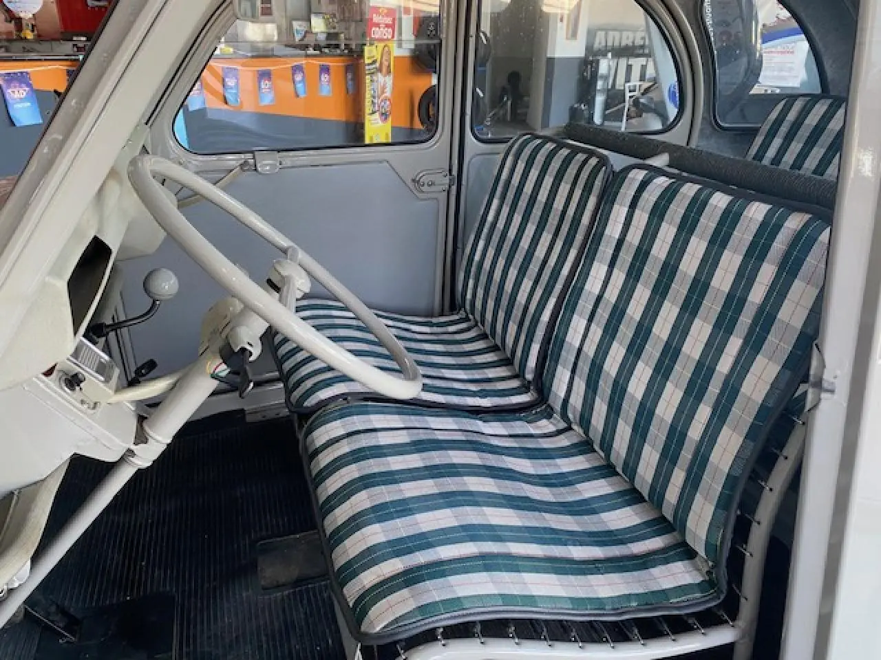 Intérieur de la Citroën 2 CV beige clair de 1968, vue côté conducteur montrant le volant et les sièges à motifs rayés.