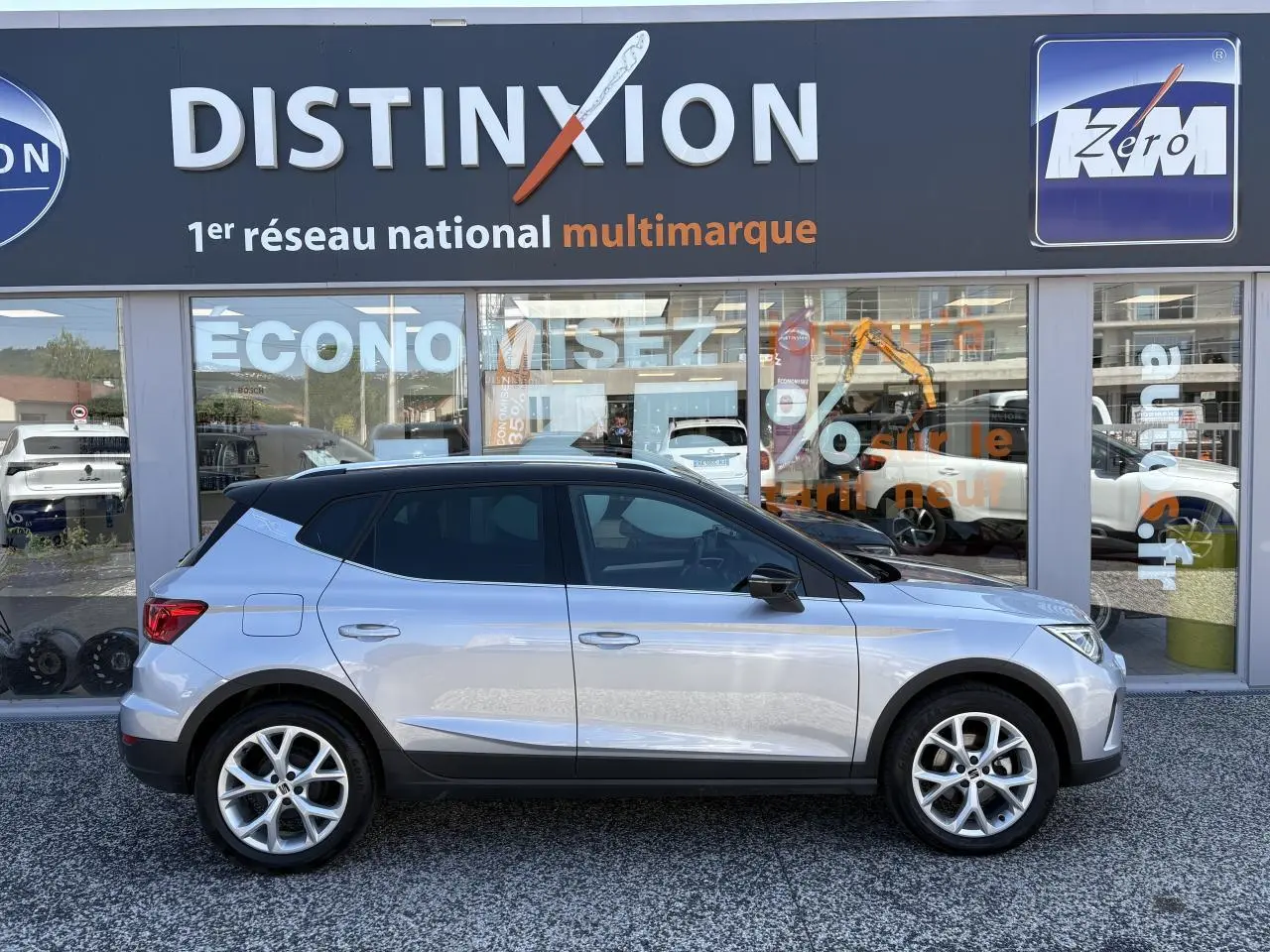 Vue de profil côté gauche d'un SEAT Arona 2024 gris clair avec toit noir et jantes alliage 16 pouces devant un showroom.