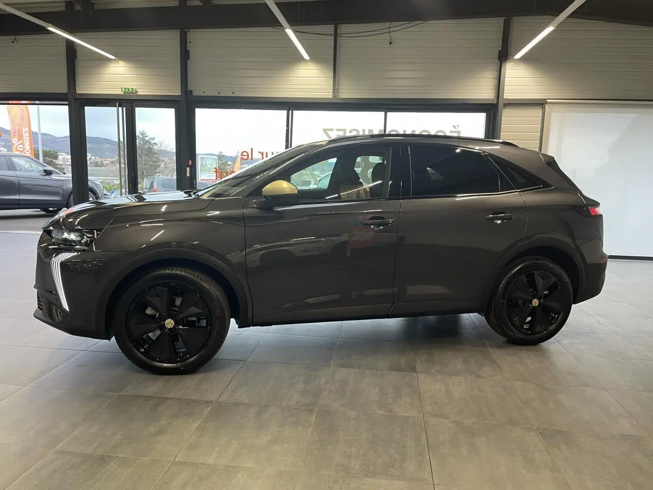 Vue de profil côté gauche du DS7 Crossback gris titanium avec jantes noires et coques de rétroviseurs dorées satinées en intérieur showroom.