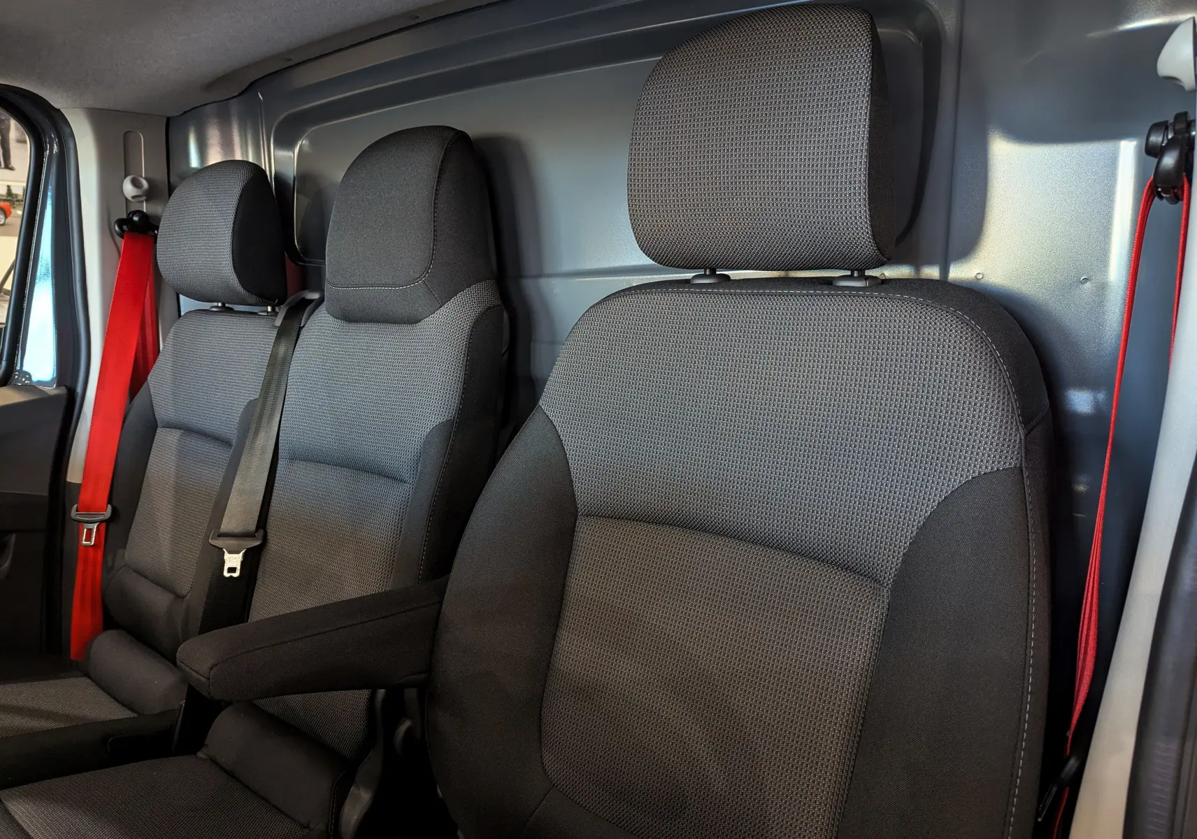Vue intérieure rapprochée des sièges avant en tissu gris foncé du Renault Trafic Fourgon L2H1 2025 avec ceintures rouges.
