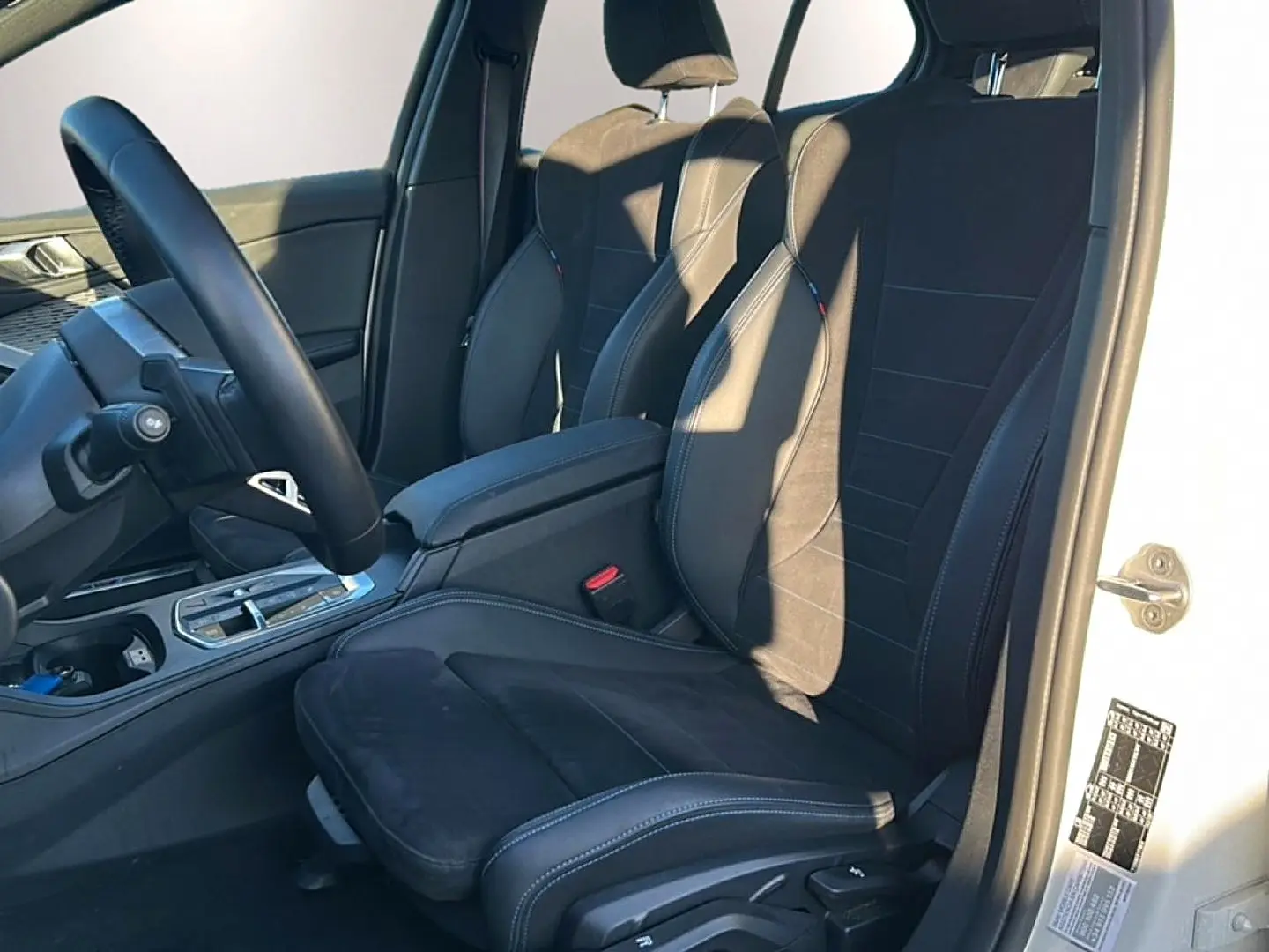 Vue intérieure côté conducteur d'une BMW Série 1 blanche, sièges sport en alcantara noir avec coutures bleues.