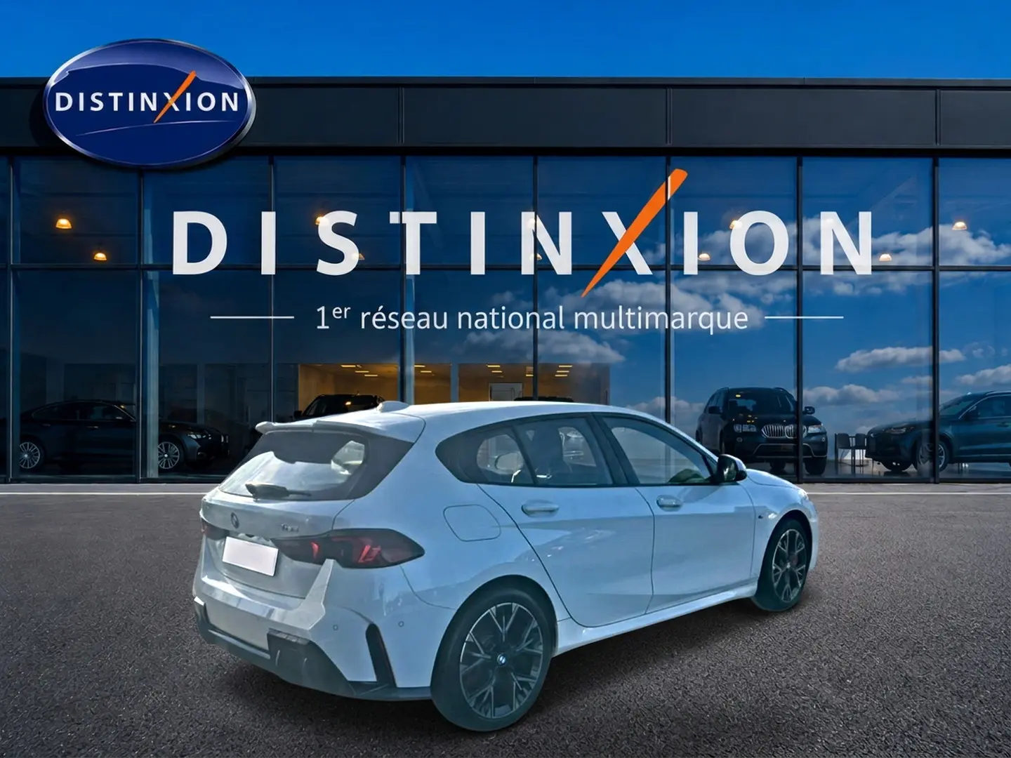 BMW Série 1 blanc alpin vue 3/4 arrière droit, avec jantes bicolores et becquet sport devant un showroom Distinxion.