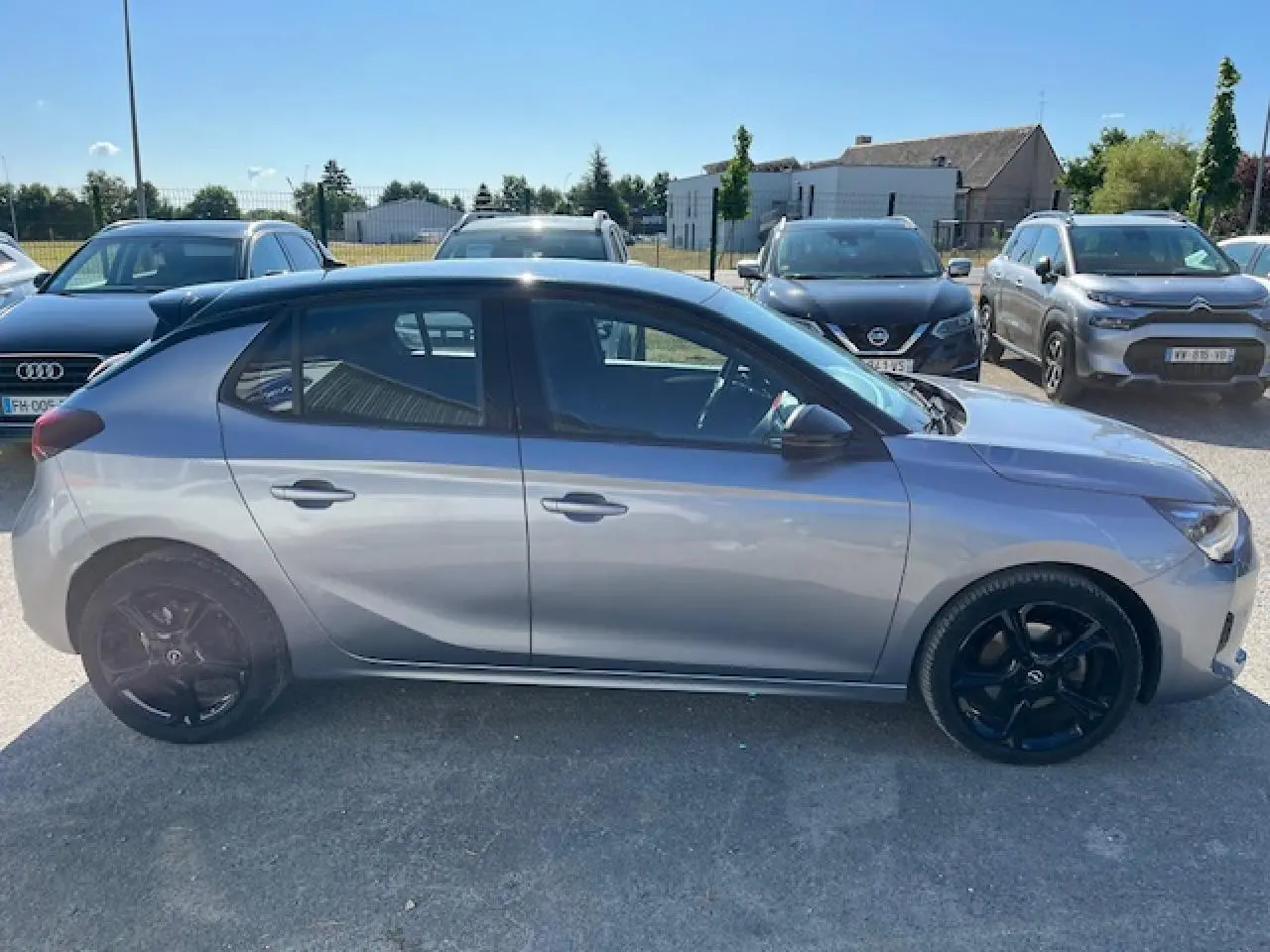 Profil côté gauche d'une Opel Corsa gris métallisé avec toit noir et jantes noires, stationnée en extérieur au soleil.