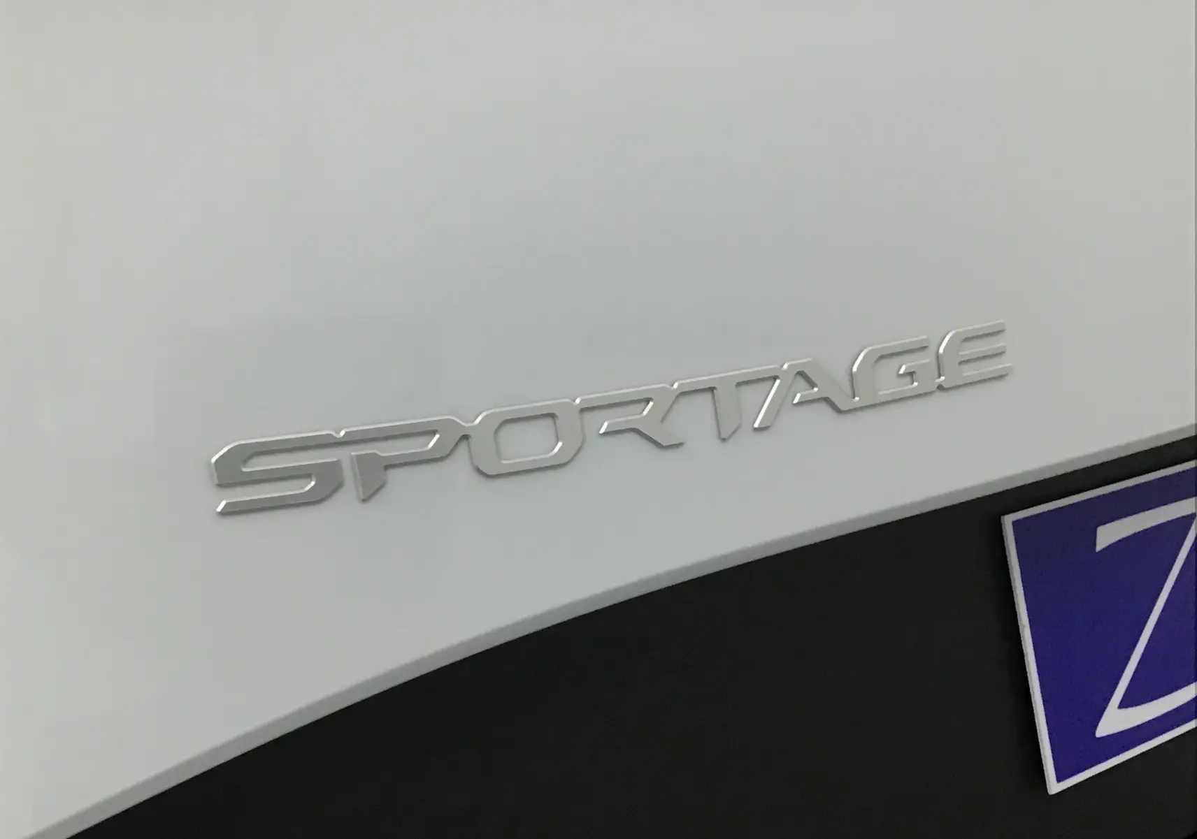 Gros plan sur l’inscription SPORTAGE en chrome sur la carrosserie blanche du SUV KIA, près du pare-chocs noir.