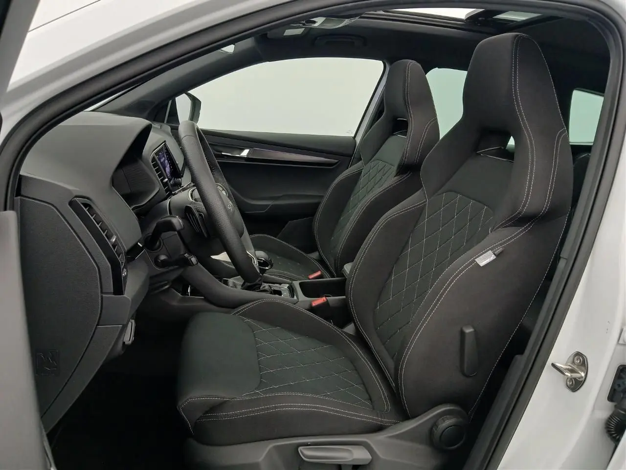 Vue intérieure côté conducteur du Skoda Karoq blanc lune 2025, sièges sport noirs avec surpiqûres blanches.