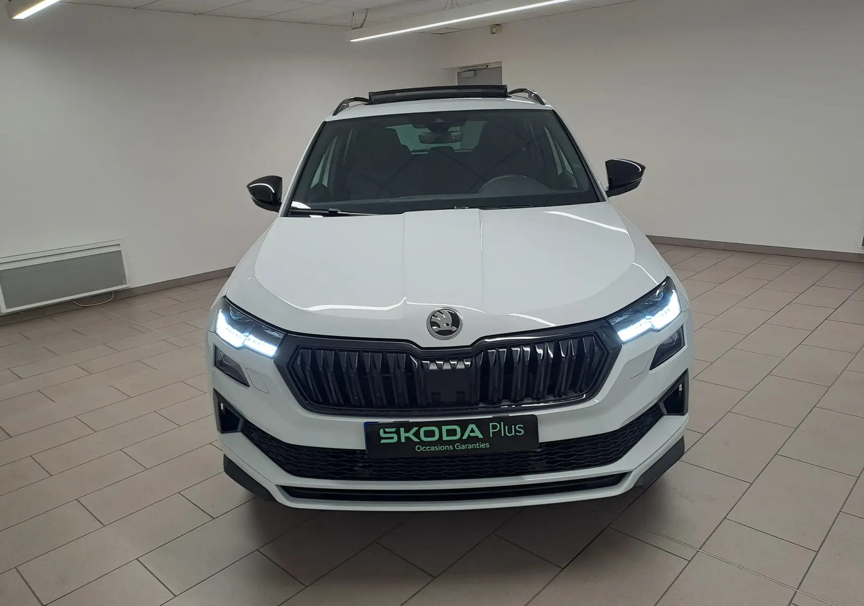 Vue avant d'un Skoda Karoq blanc lune 2025 avec phares allumés et toit ouvrant panoramique visible en intérieur.