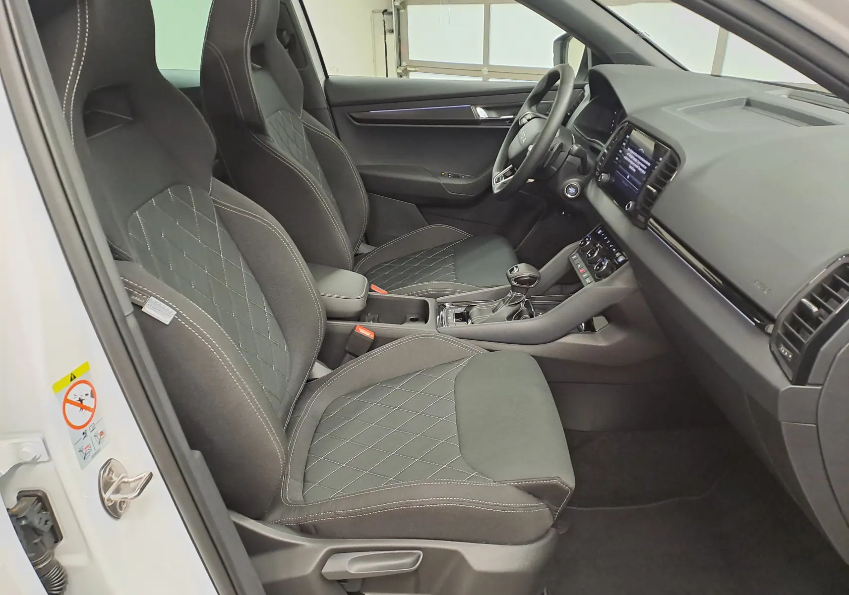 Vue intérieure côté conducteur de la Skoda Karoq blanc lune 2025, sièges sport noirs avec surpiqûres contrastées.