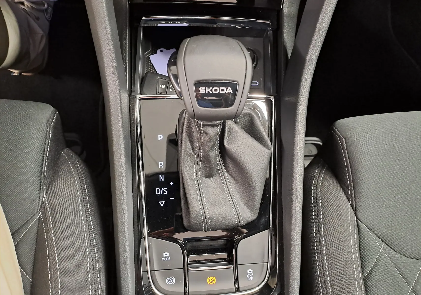 Vue plongeante sur le levier de vitesse automatique noir marqué SKODA, entouré de sièges tissu gris avec surpiqûres blanches.