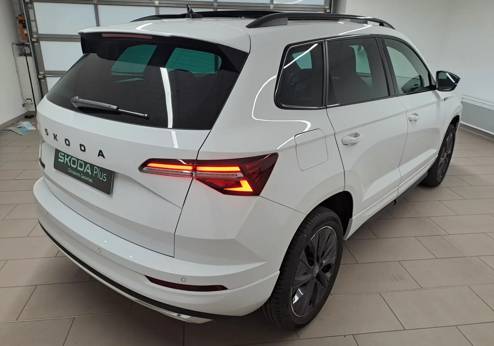 Vue 3/4 arrière droite du Skoda Karoq blanc Lune avec feux arrière allumés et toit panoramique fermé.