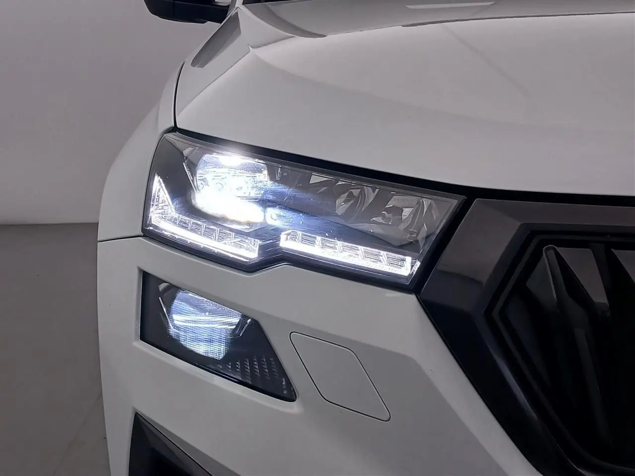 Gros plan sur le phare avant gauche allumé d’un Skoda Karoq blanc lune 2025 avec calandre noire sportive.