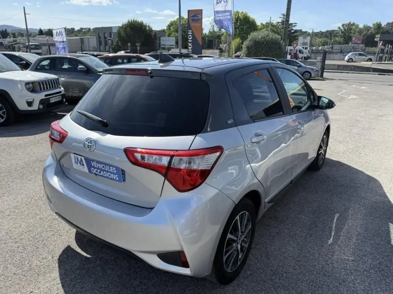 Toyota Yaris gris métal vue 3/4 arrière droit, avec vitres teintées et feux arrière distinctifs, en extérieur sur parking.