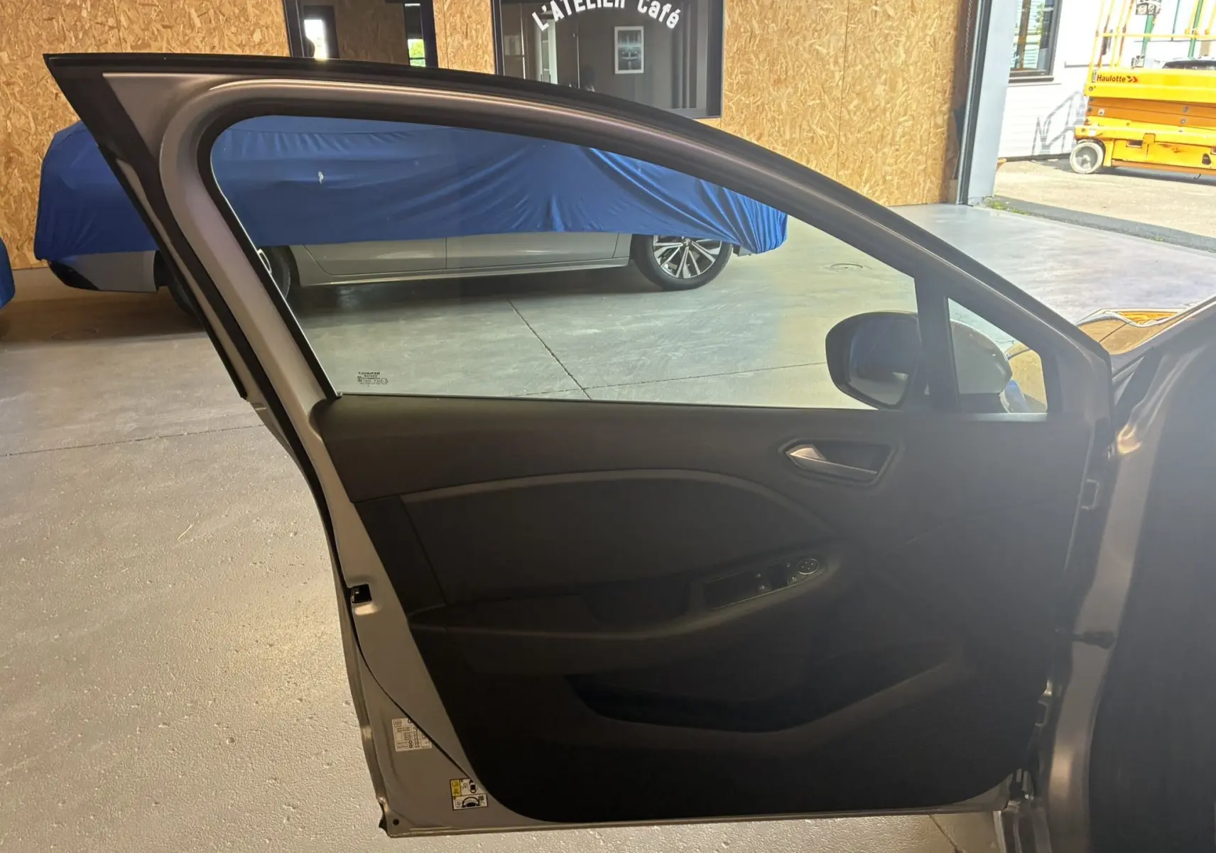 Vue intérieure de la porte avant gauche ouverte d'une Renault Clio 1.5 BlueDCI Business grise, montrant la garniture noire et les commandes.