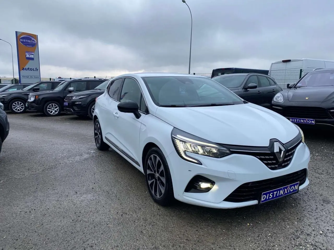 Renault Clio blanche vue 3/4 avant droit avec jantes alliage noires et feux LED allumés sur parking.
