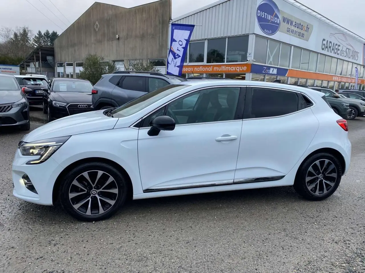 Profil côté gauche d'une Renault Clio blanche 2023 avec jantes alliage noires et vitres surteintées, stationnée en extérieur.