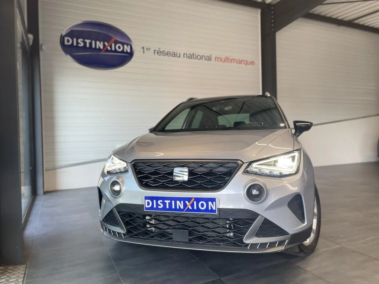 Vue avant d'un SEAT Arona 2024 gris clair avec calandre noire et phares allumés en showroom.