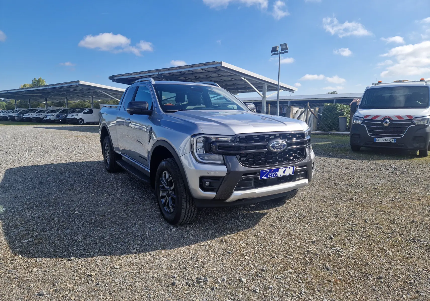 Ford Ranger Super Cabine gris Iconic vu en 3/4 avant droit sur un parking extérieur sous un ciel bleu.