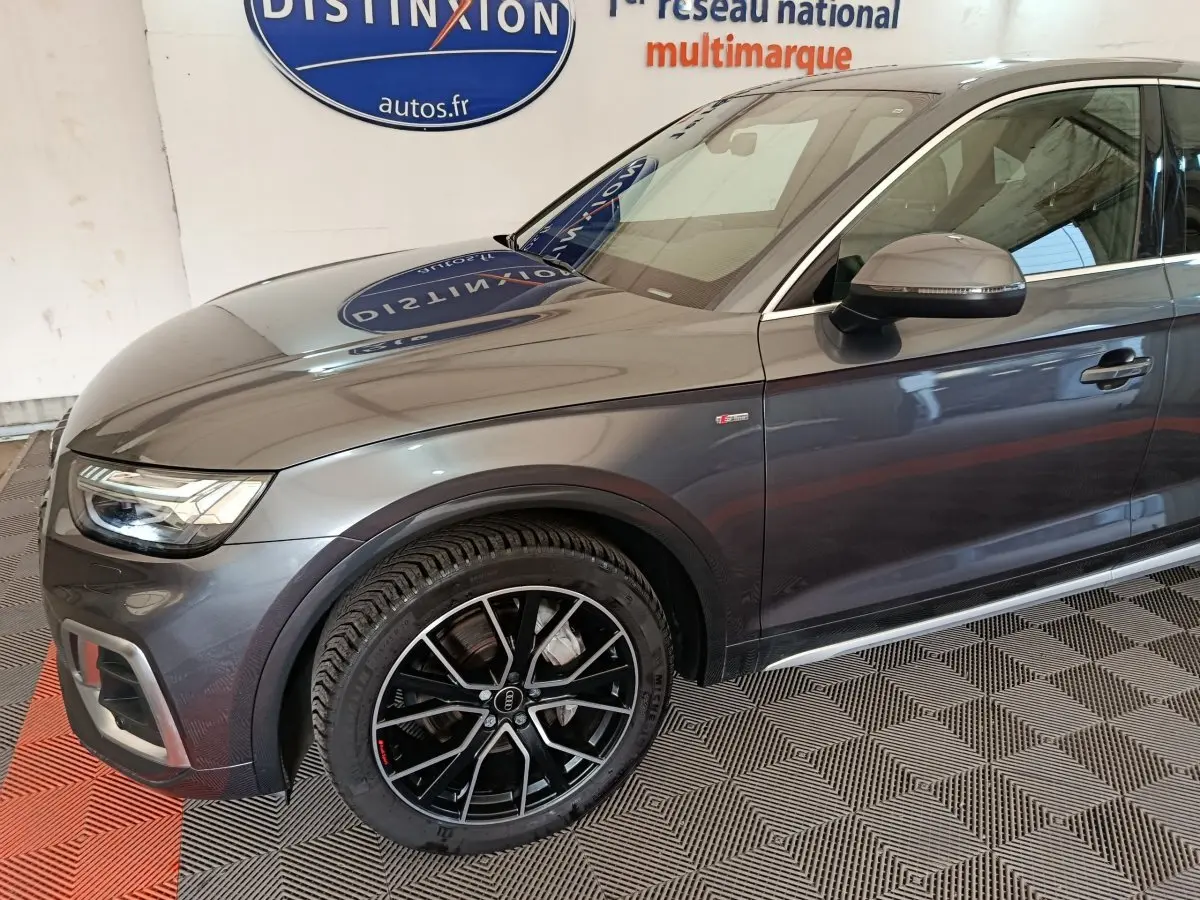 Vue de profil avant droit d'un Audi Q5 gris clair 2023 avec jantes noires et badge S line sur l'aile avant.
