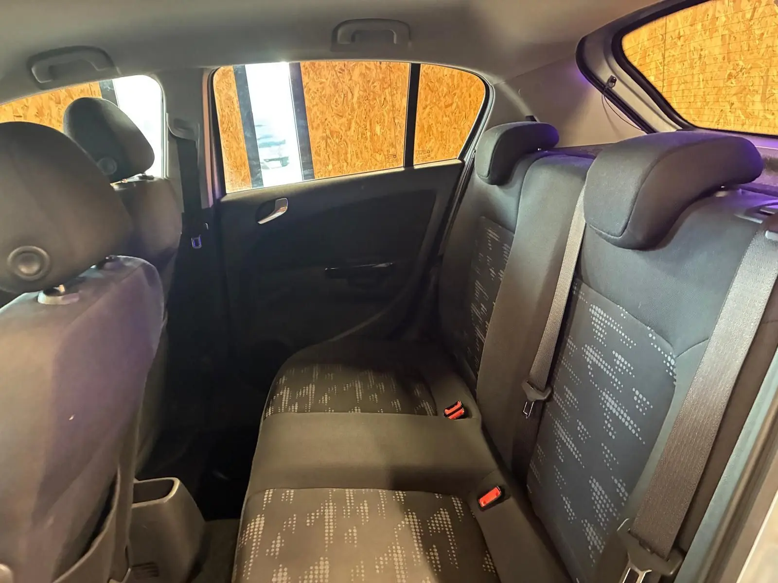 Vue intérieure arrière côté droit de l’Opel Corsa gris clair 2012, sièges tissu noir avec motifs et ceintures visibles.