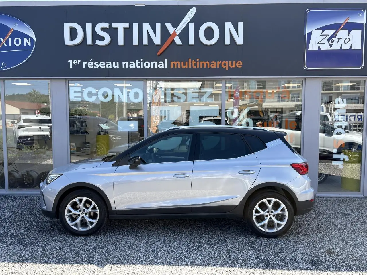 Vue latérale droite du SEAT Arona 2024 en gris clair avec toit noir et jantes alliage 16 pouces devant un showroom.