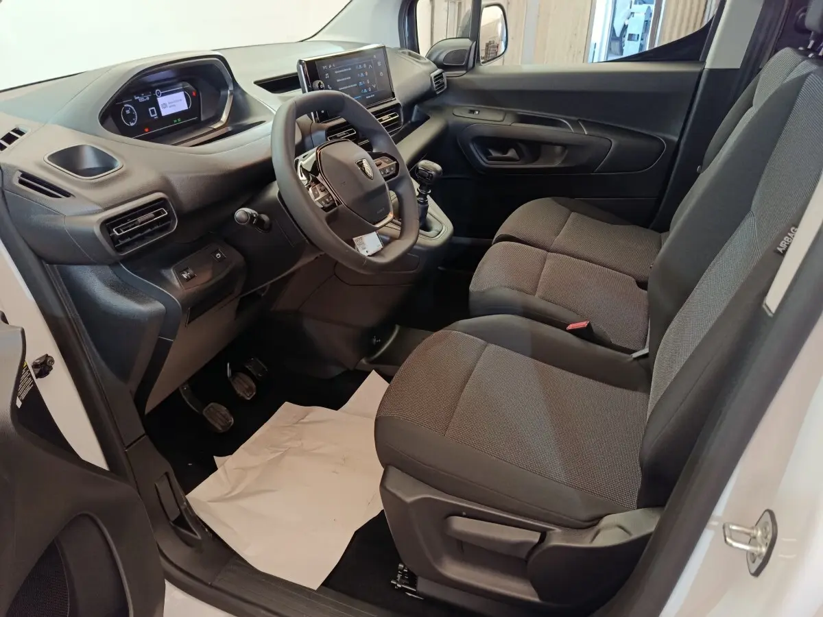 Vue intérieure côté conducteur du Peugeot Partner blanc 2025, montrant le tableau de bord, volant et sièges tissu gris.
