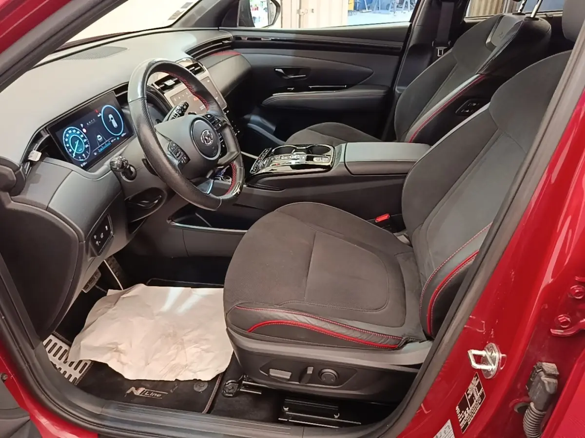 Vue intérieure côté conducteur du Hyundai Tucson rouge, avec sièges noirs à surpiqûres rouges et tableau de bord digital.