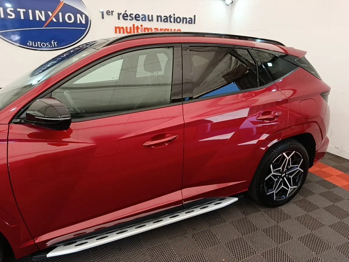 Vue latérale gauche du Hyundai Tucson rouge 2022 avec jantes noires et marchepieds argentés visibles en intérieur showroom.