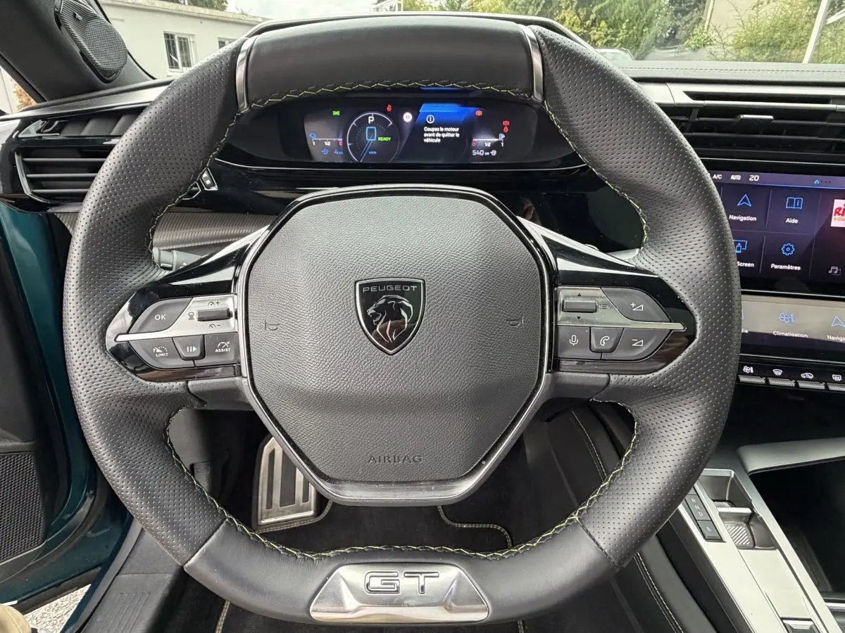 Vue rapprochée du volant cuir noir GT avec logo Peugeot et tableau de bord numérique du Peugeot 408 PHEV 225 bleu.