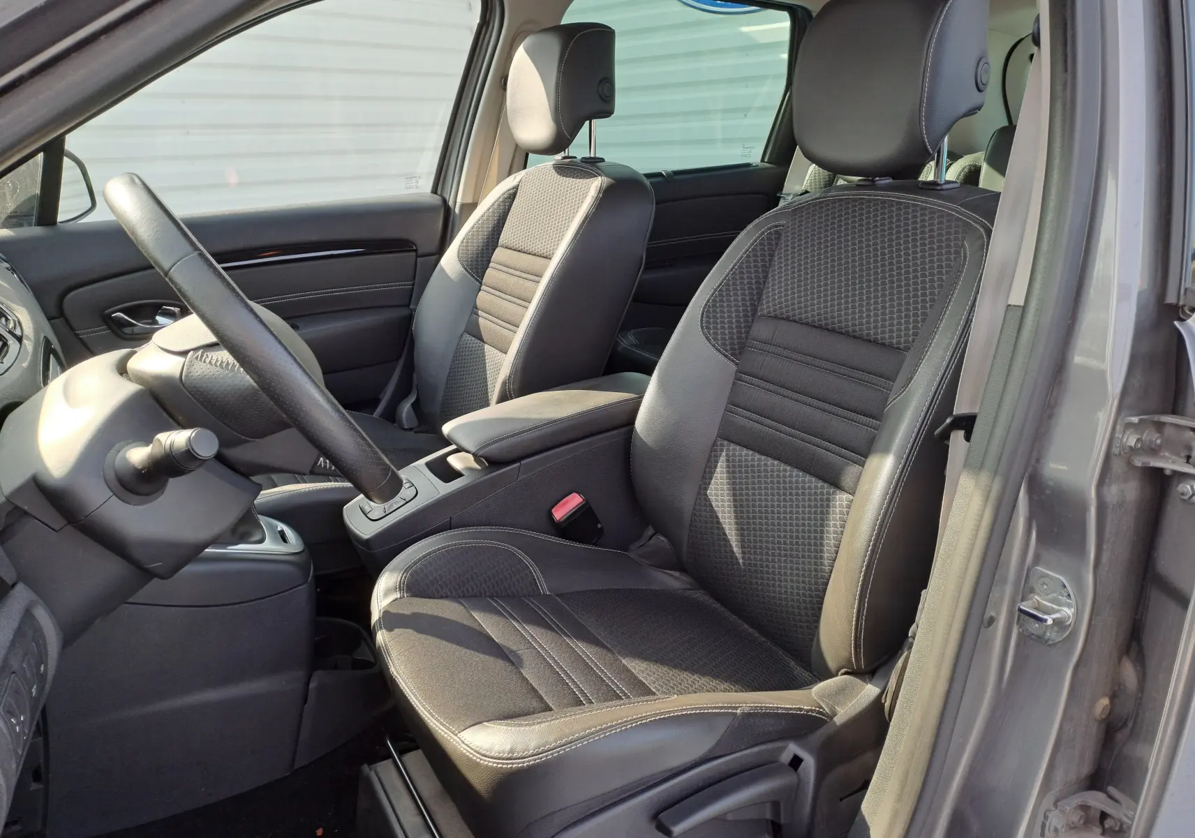 Vue intérieure côté conducteur du Renault Scenic III gris, mettant en valeur les sièges en tissu noir avec surpiqûres et le volant.