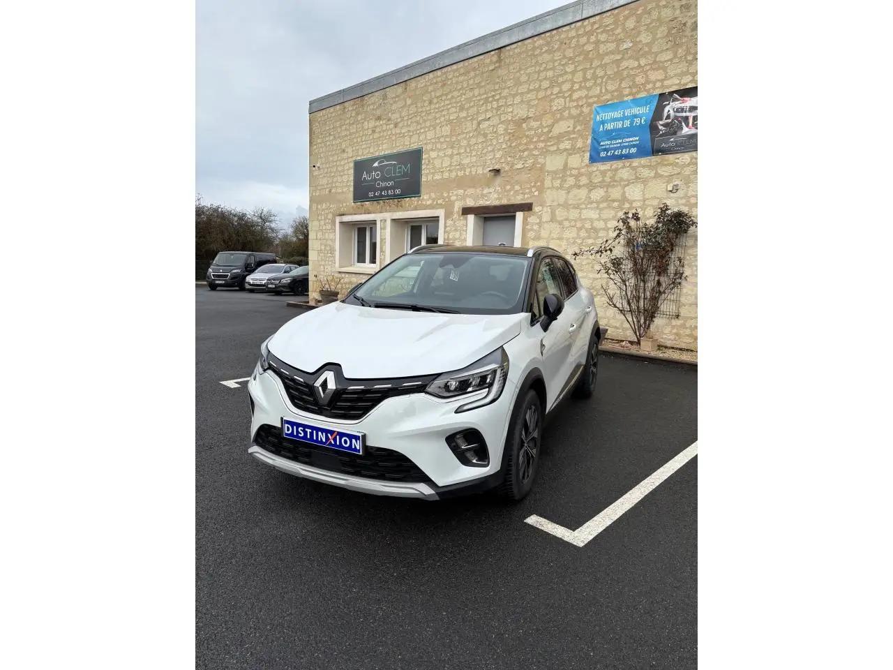 Renault Captur blanc en 3/4 avant droit, avec toit noir et calandre noire brillante, stationné devant un mur en pierre.