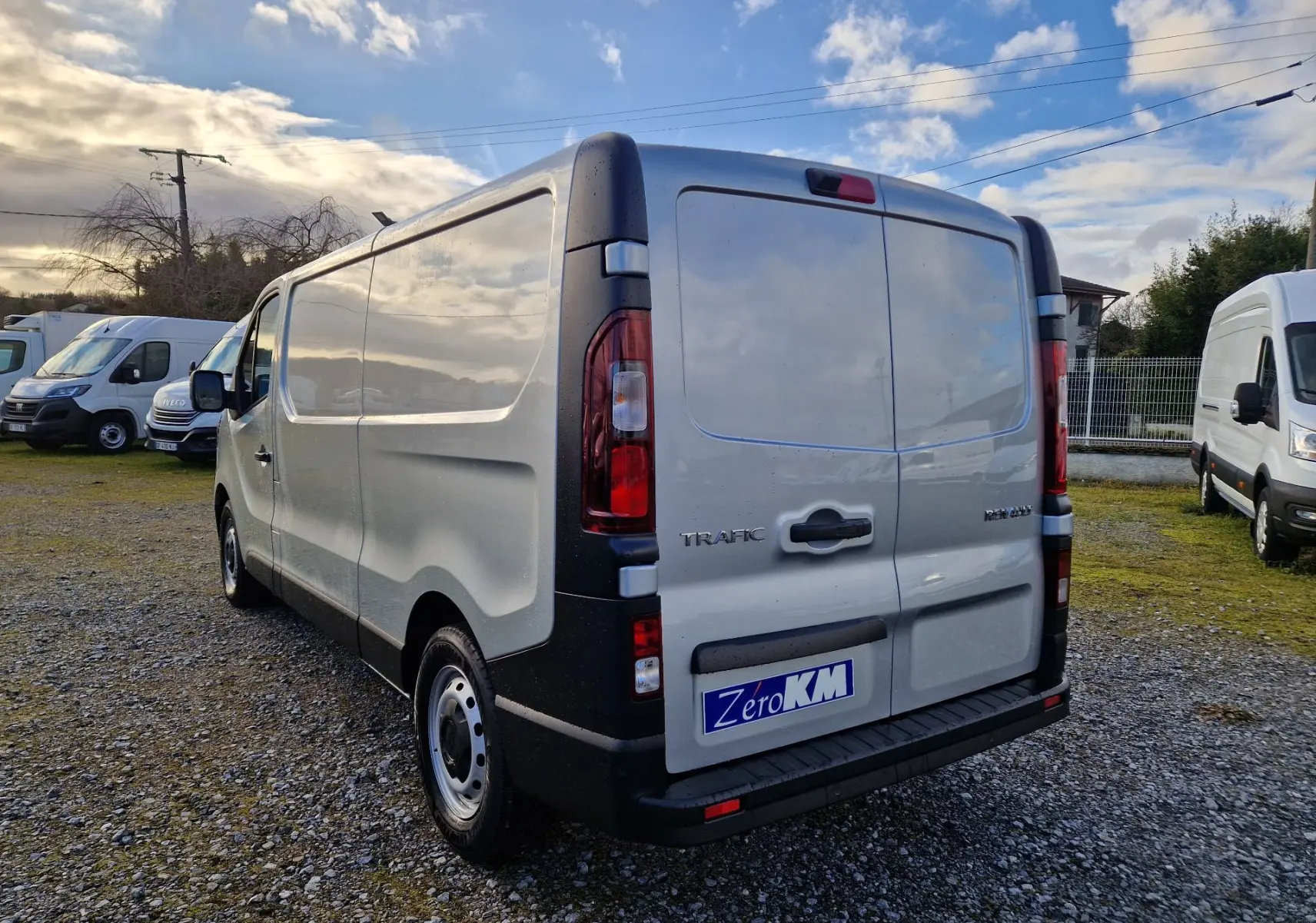Vue 3/4 arrière droite d’un Renault Trafic Fourgon gris Highland avec portes arrière fermées sur un sol gravillonné.
