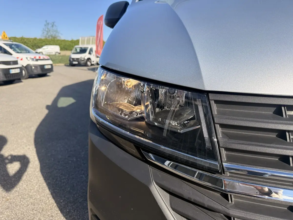 Gros plan sur le phare avant droit et la calandre d’un Volkswagen Transporter gris clair sous un ciel bleu.