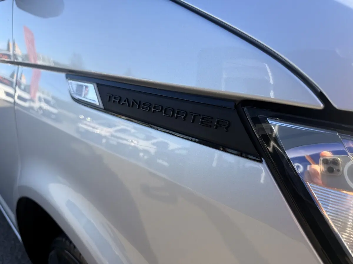 Gros plan sur l'aile avant gauche gris clair du Volkswagen Transporter avec logo noir "TRANSPORTER" et phare partiel visible.