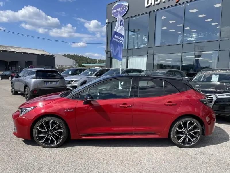 Profil côté gauche d'une Toyota Corolla 184 H Collection rouge métal avec toit noir, roues alliage, stationnée devant un concessionnaire.