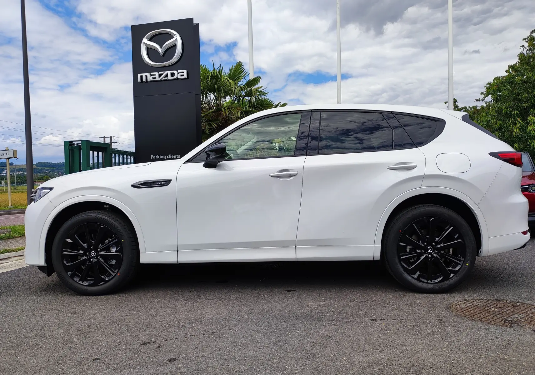 Vue latérale droite d'un Mazda CX-60 blanc métal 2024 avec jantes noires et vitres teintées, devant un panneau Mazda.