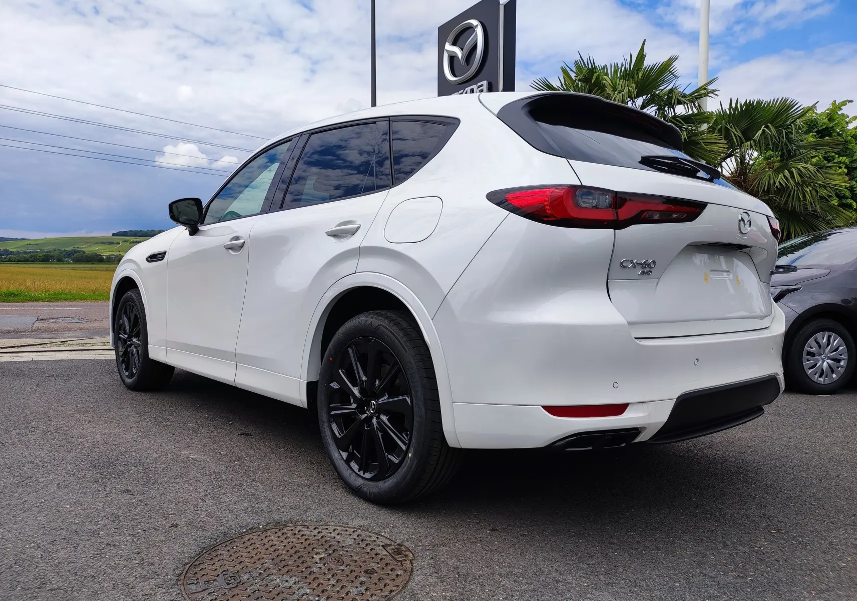Vue 3/4 arrière gauche d'un Mazda CX-60 blanc métal avec jantes noires et vitres teintées, stationné devant un concessionnaire.