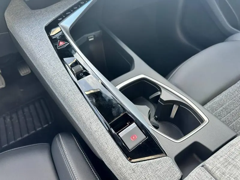 Gros plan sur la console centrale grise et noire du Peugeot 3008 Hybrid 2025, avec porte-gobelets et bouton frein à main électronique.