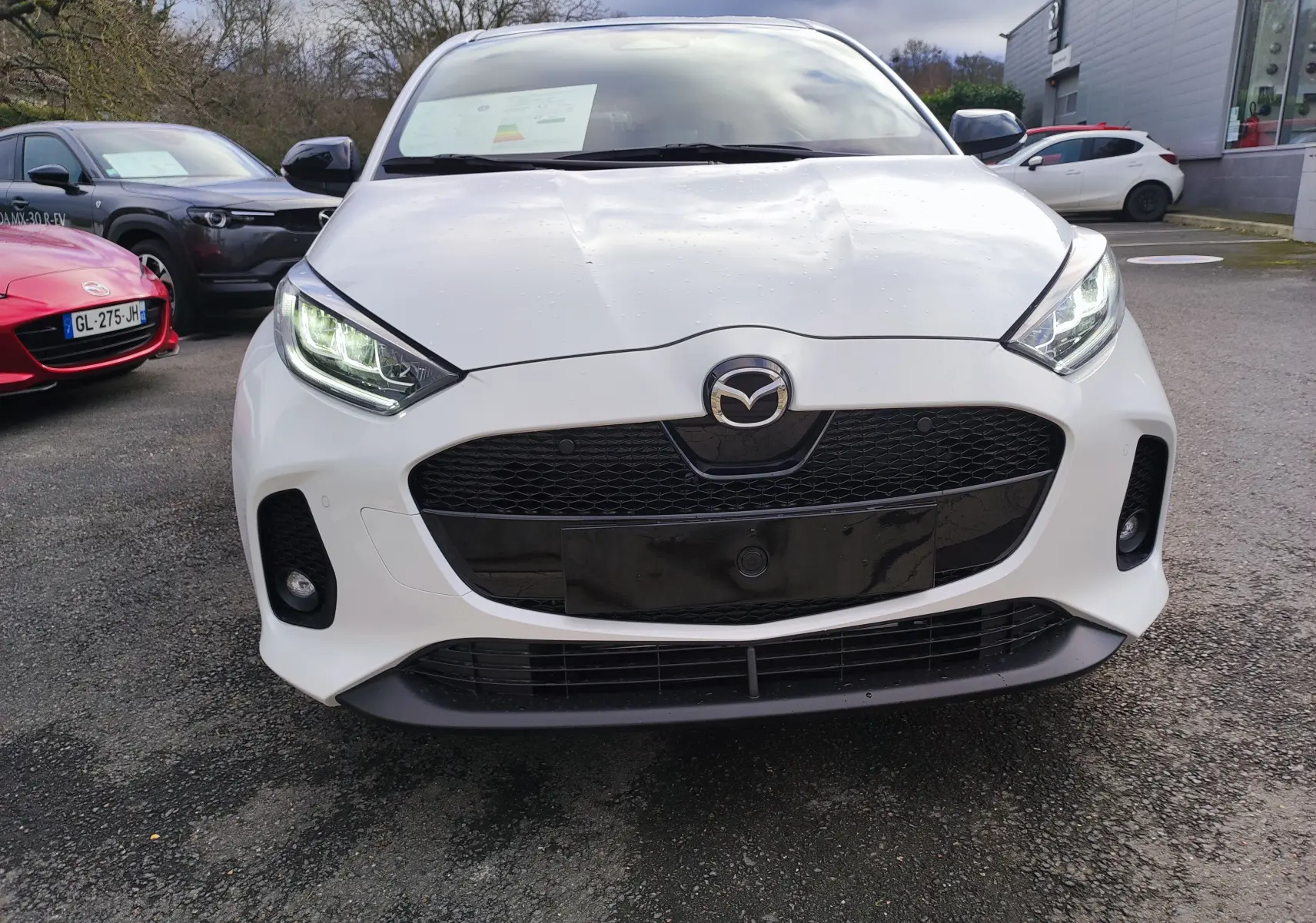 Vue frontale d'une Mazda2 Hybrid 2024 blanc métallisé avec calandre noire et phares LED allumés.