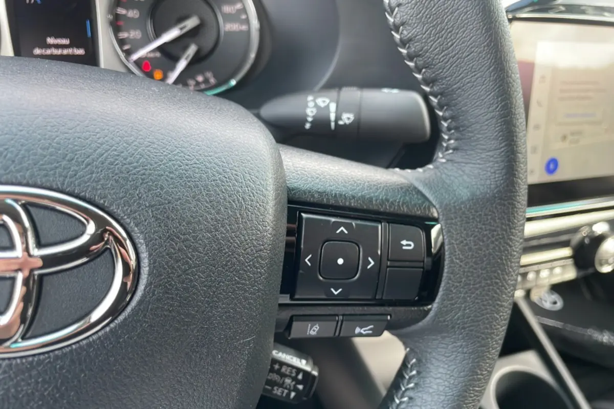 Gros plan sur le volant en cuir noir du Toyota Hilux 4x4 2025, avec commandes multifonctions et logo Toyota visible.