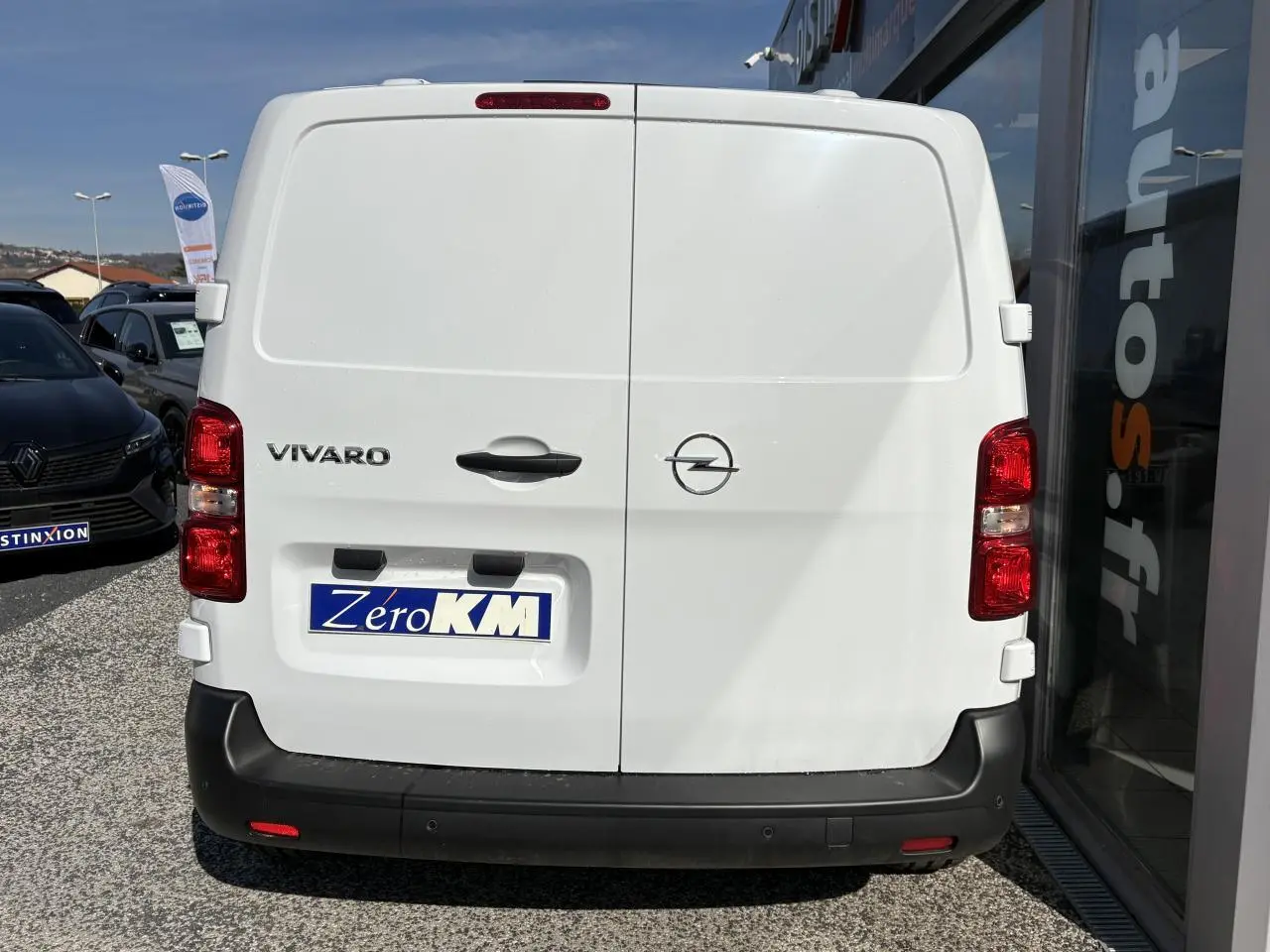 Vue arrière d’un utilitaire Opel Vivaro blanc avec portes battantes et pare-chocs noir, stationné près d’un bâtiment vitré.