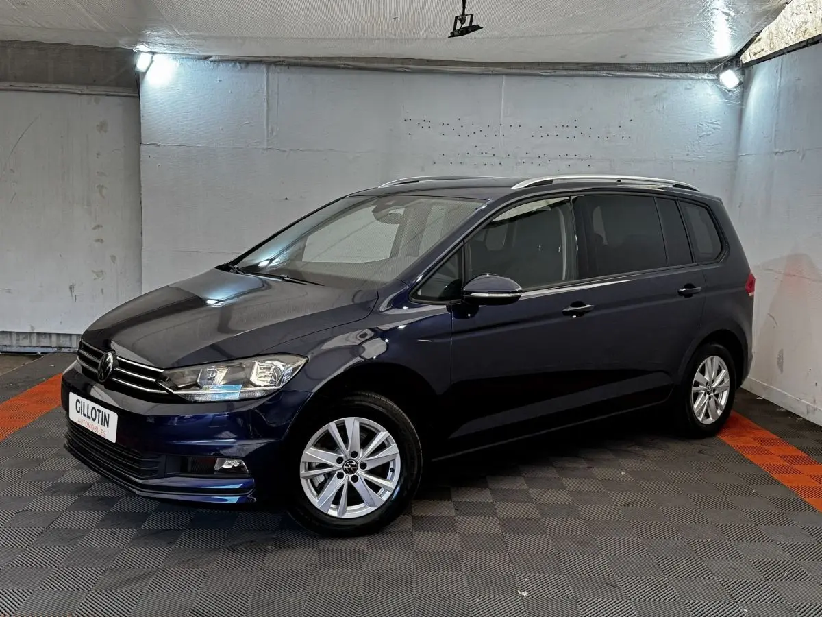 Volkswagen Touran bleu 3/4 avant droit en intérieur, avec jantes alliage et barres de toit chromées.