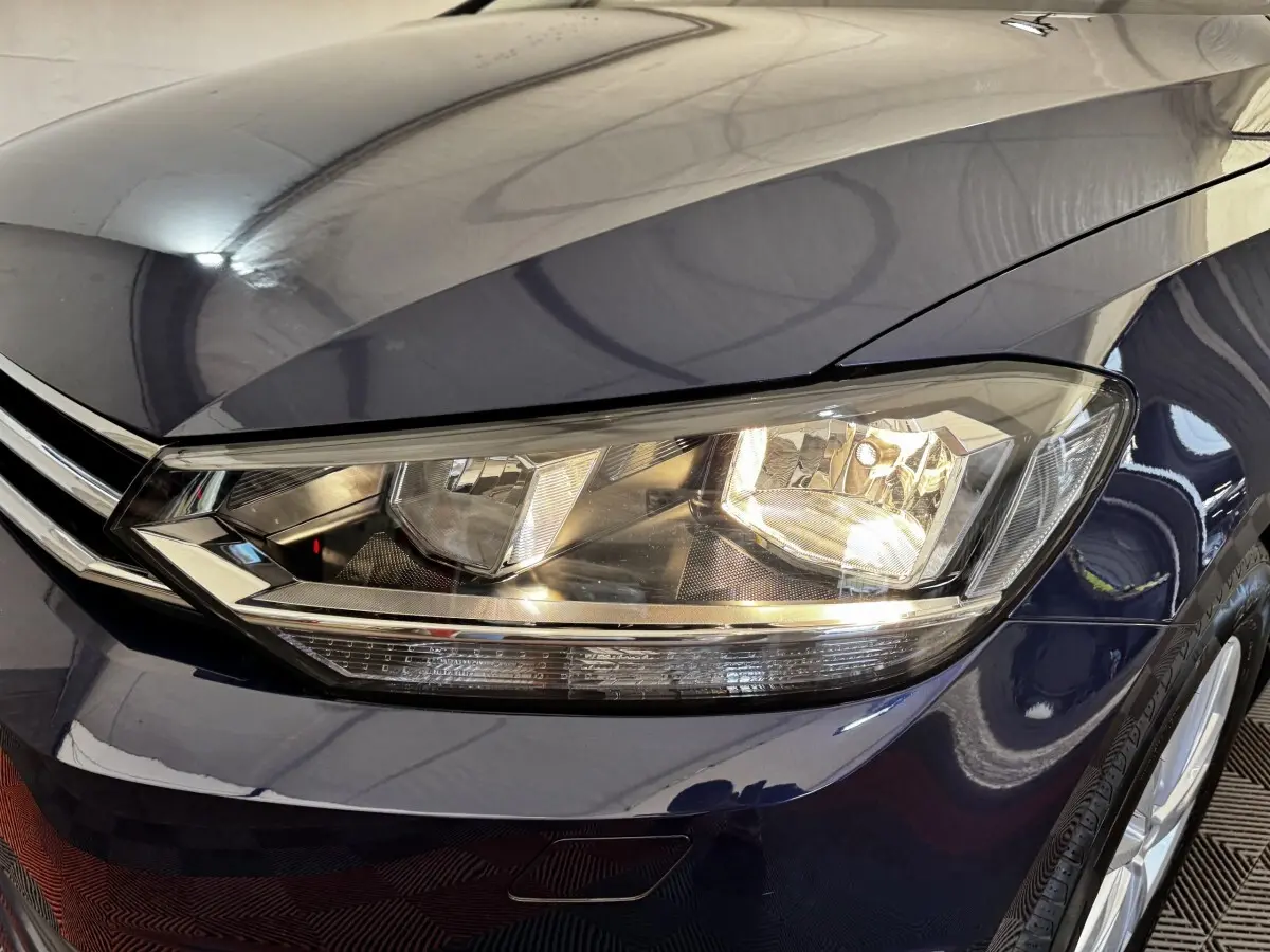Gros plan sur le phare avant droit allumé d’un Volkswagen Touran bleu 2024, avec partie de la calandre visible.