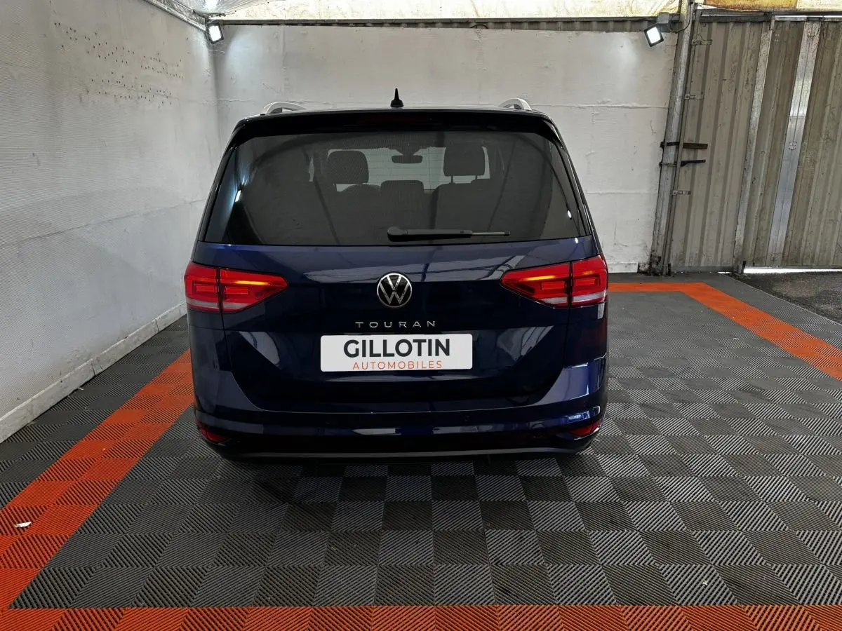 Vue arrière d'un Volkswagen Touran bleu 2024 garé dans un garage avec sol à damier noir et orange.