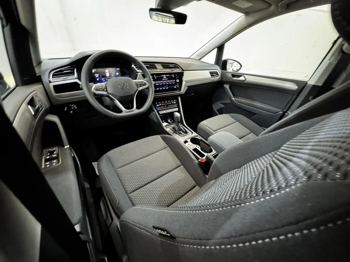 Vue intérieure côté conducteur du Volkswagen Touran 2024, montrant tableau de bord et sièges tissu gris foncé.