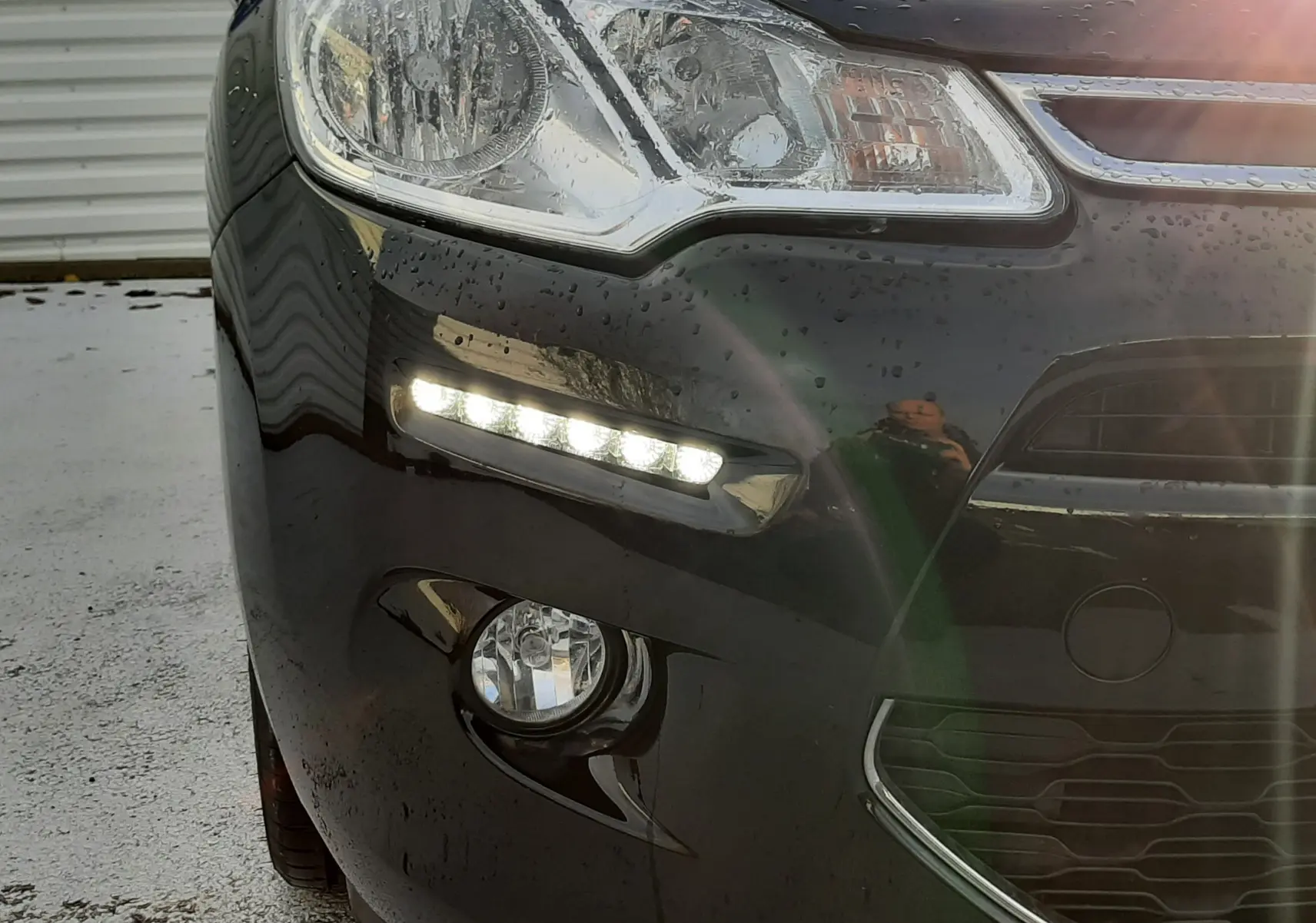 Gros plan sur l'avant gauche noir d'une Citroën C3 2016 avec feux diurnes LED allumés et carrosserie mouillée.