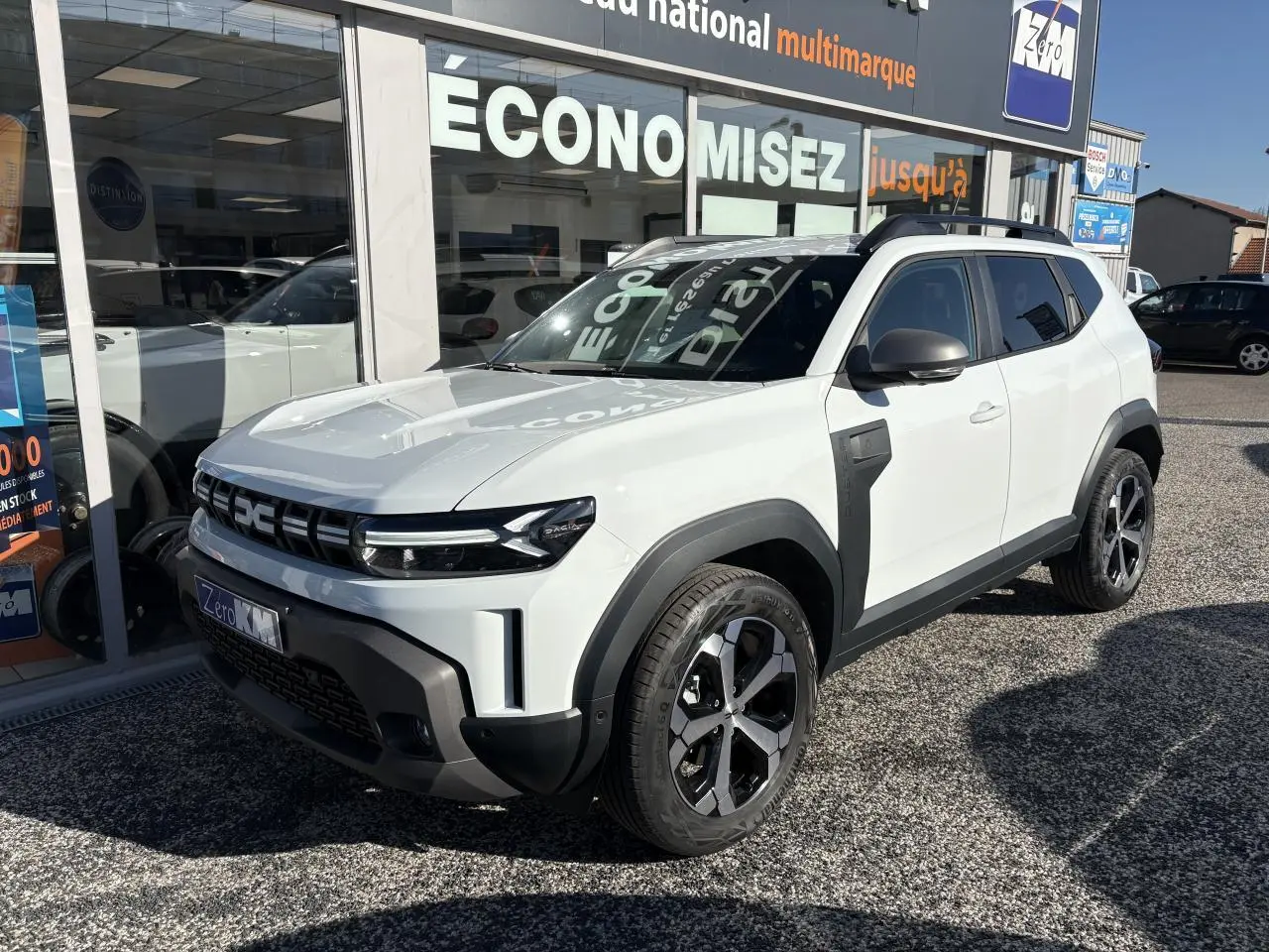 SUV Dacia Duster blanc métallisé en 3/4 avant droit, avec barres de toit noires et jantes alliage 18 pouces.