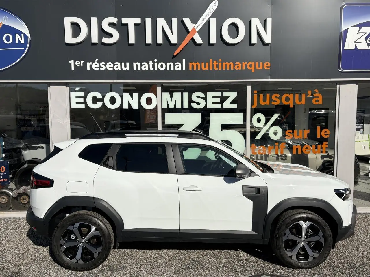 Profil droit du Dacia Duster 1.6 Hybrid blanc métallisé avec jantes alliage noires devant une vitrine commerciale.