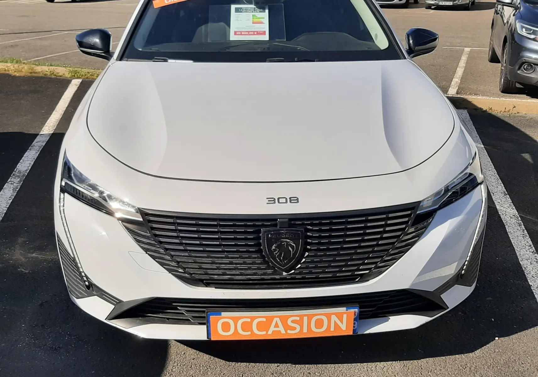 Vue avant d'une Peugeot 308 blanche 2024 avec calandre noire et plaque "OCCASION" en orange, en extérieur.