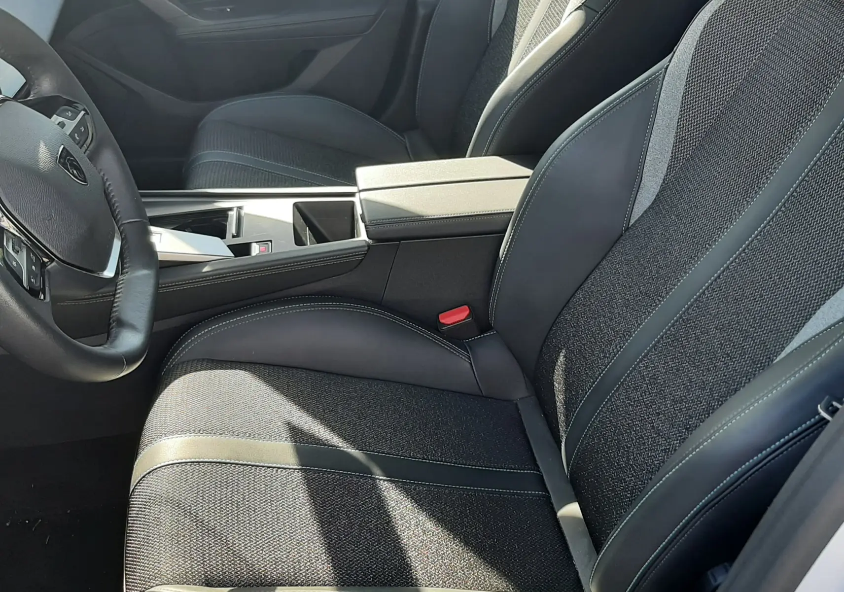 Vue intérieure côté conducteur de la Peugeot 308 2024, sièges tissu gris avec surpiqûres blanches et console centrale noire.
