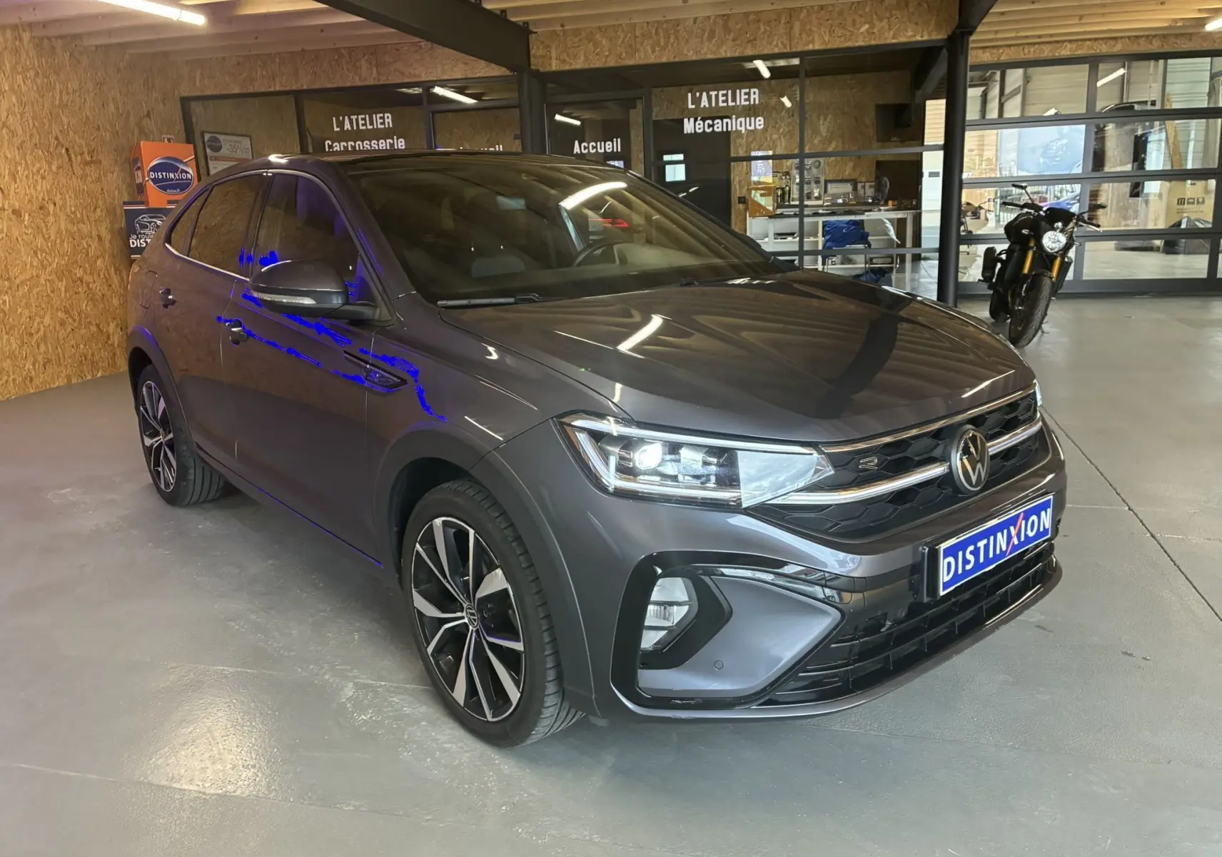 Volkswagen Taigo 1.5 TSI R-Line gris vu en 3/4 avant droit dans un atelier avec éclairage intérieur.