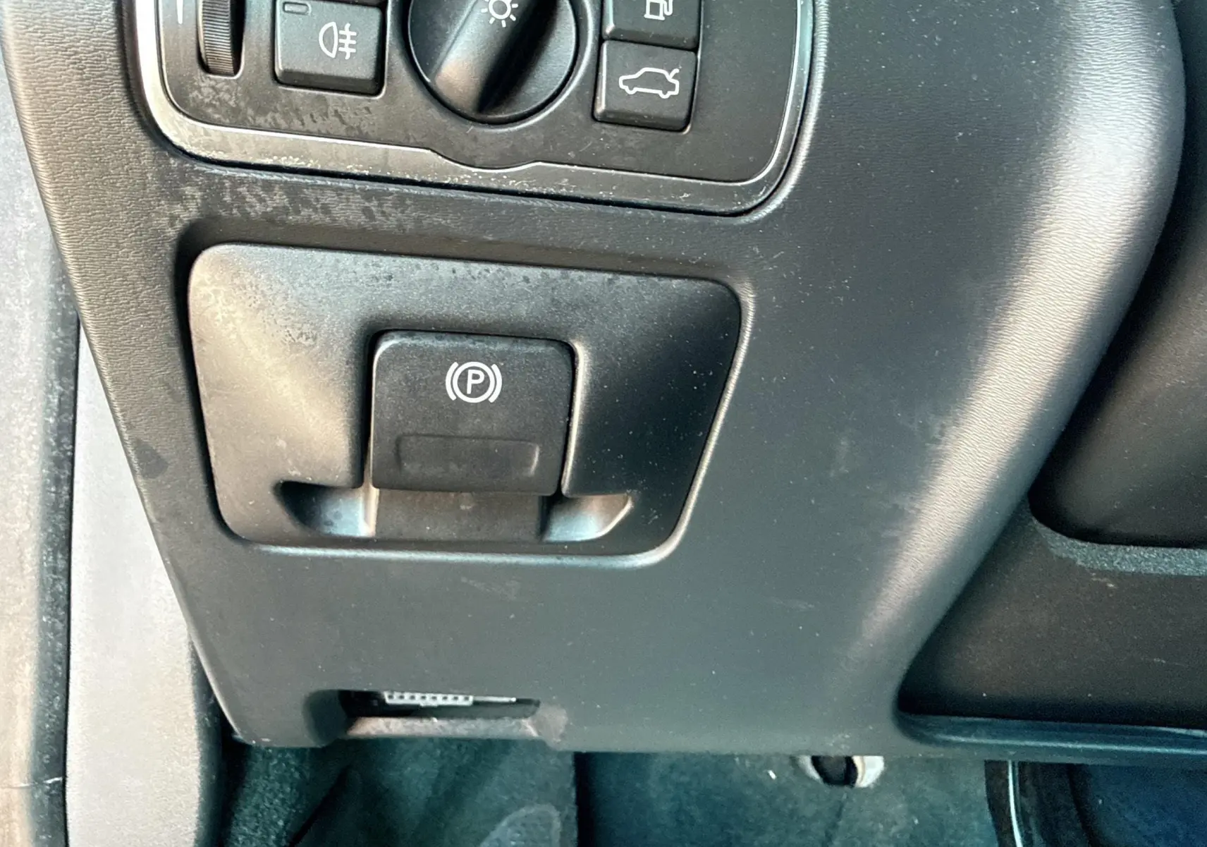 Gros plan sur le bouton de frein à main électrique et commandes d’éclairage du tableau de bord d’un Volvo XC60 gris 2016.